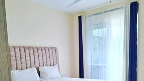 Ein ordentlich bezogenes Bett mit Kopfteil, ein Fenster mit Vorhängen und ein Deckenventilator.