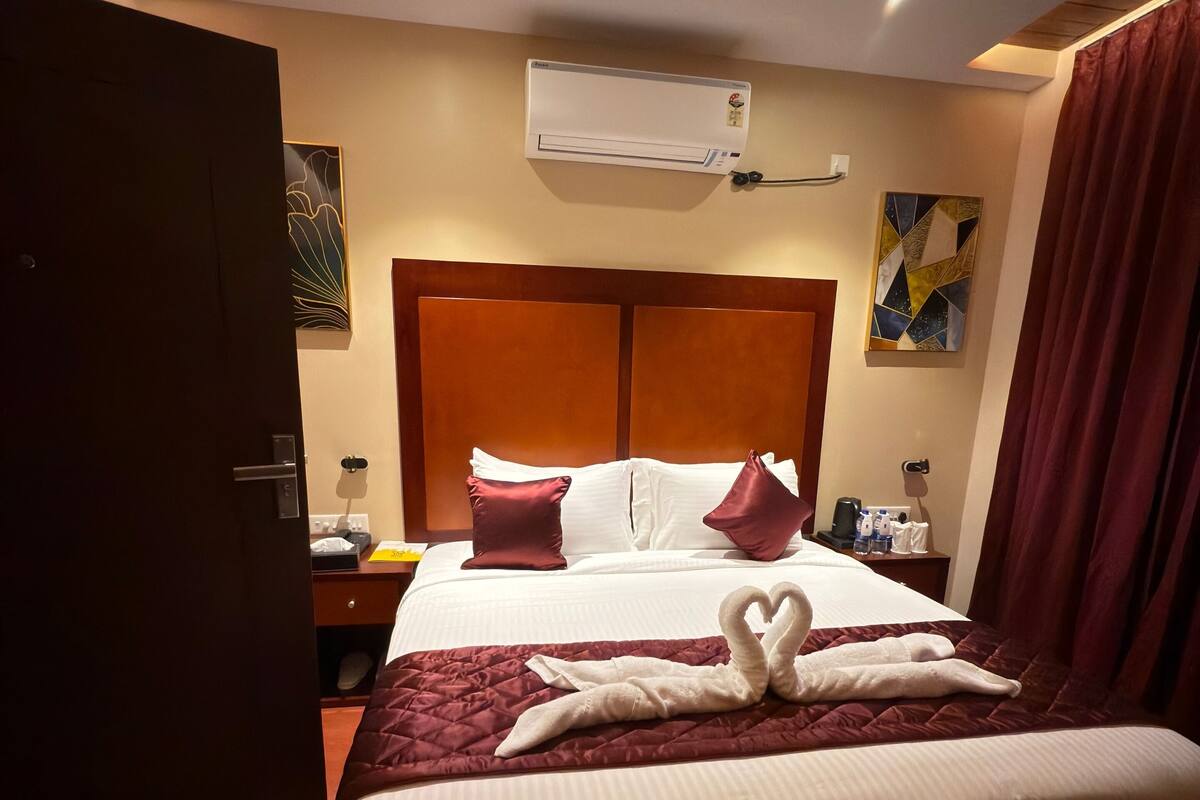 Een hotelkamer met een groot bed, een houten hoofdbord, een airconditioningapparaat, een nachtkastje met een lamp en een schilderij aan de muur.