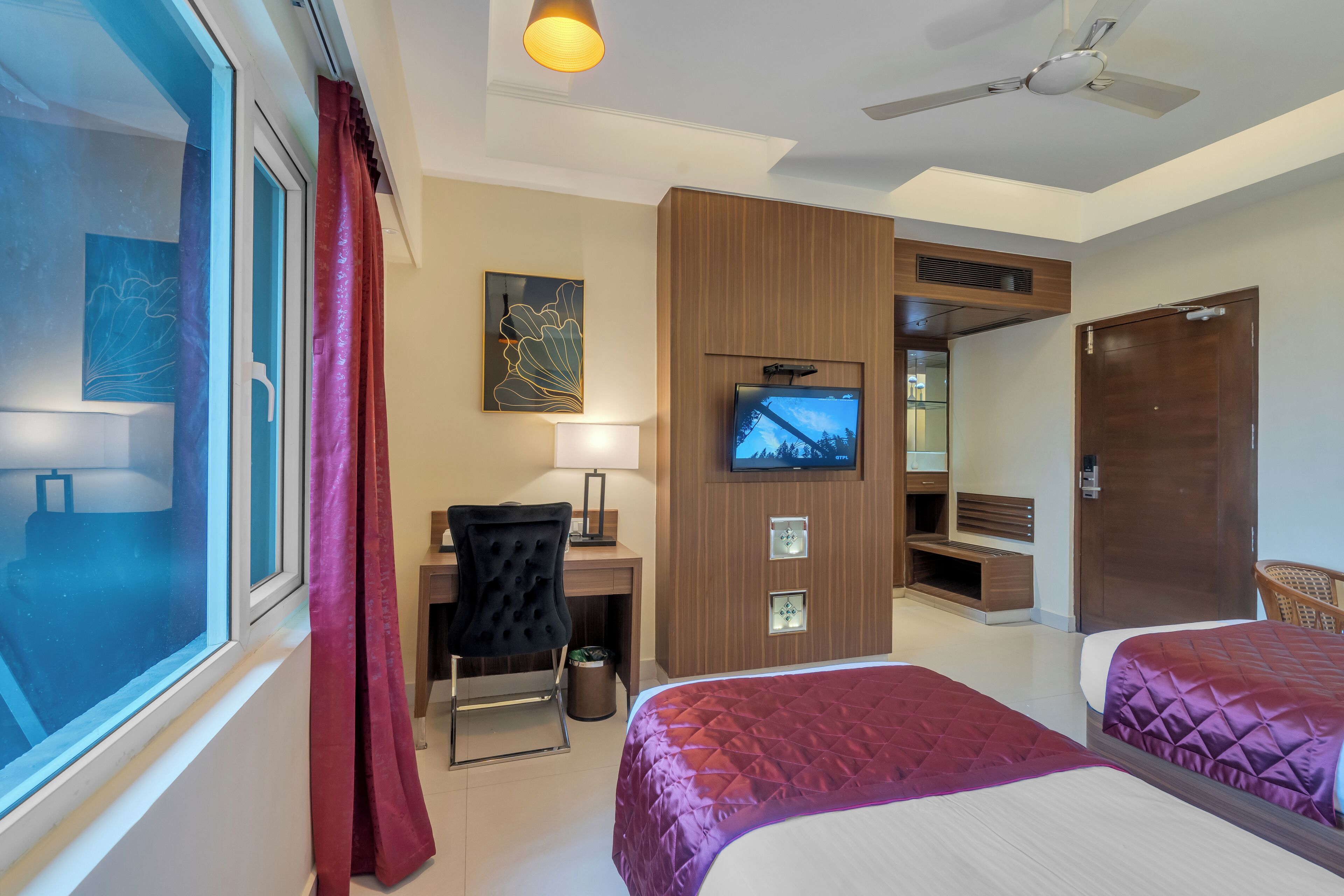 A hotel room with two beds, a desk with a chair, a TV, and a ceiling fan.