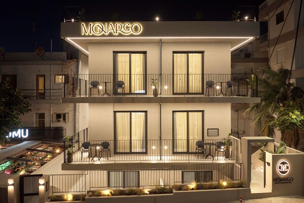 Un hôtel moderne, reconnaissable à son grand panneau « MONARCO », doté de balcons agrémentés de sièges et de plantes en pot, et d’une entrée bien éclairée.