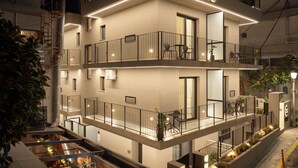 A modern multi-story building with balconies, outdoor seating, and a well-lit facade. - Monarco Executive Rooms (Rhodes)