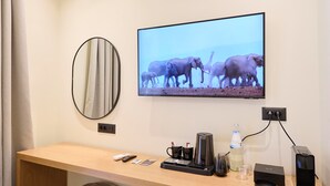 A hotel room with a wooden desk, a flat-screen TV displaying elephants, a coffee maker, and a bottle of water. - Monarco Executive Rooms (Rhodes)