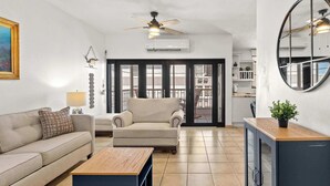 Un salon moderne avec un canapé, une table basse et un téléviseur. On y trouve également un ventilateur de plafond, un grand miroir et une plante en pot.