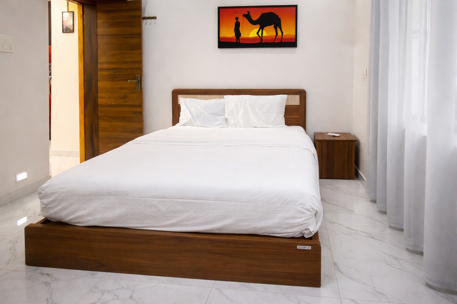 A bed with white bedding, a wooden headboard, a nightstand, a remote control, and a wall-mounted picture of a camel and a person.