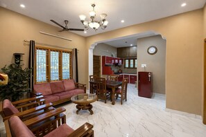 A living room with a sofa, coffee table, and dining area with chairs. There is a ceiling fan, a chandelier, and a kitchen visible through an archway. - SRI APARTMENT HOMESTAY 2BHK (Puducherry)
