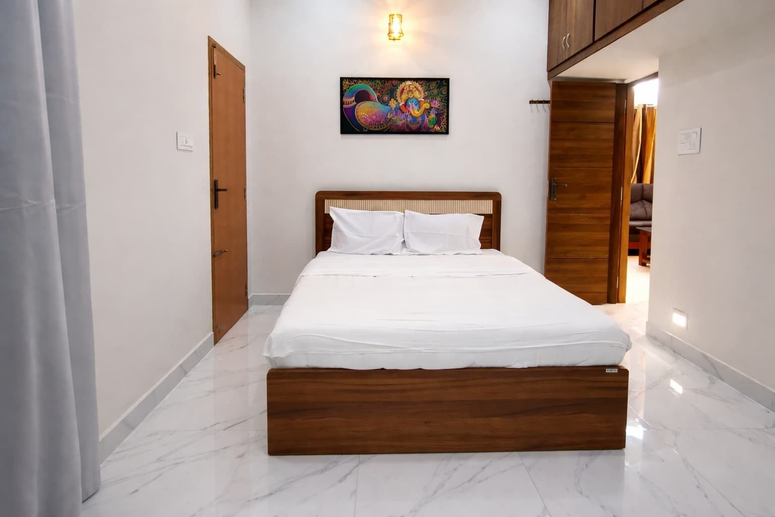 A bedroom with a wooden bed, white bedding, a colorful painting on the wall, and a wooden wardrobe.