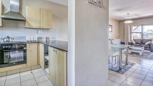 A modern kitchen with a stove, oven, and cabinets, and a living area with a sofa and a dining table. - Hideout Hotel Apartment (Sandton)