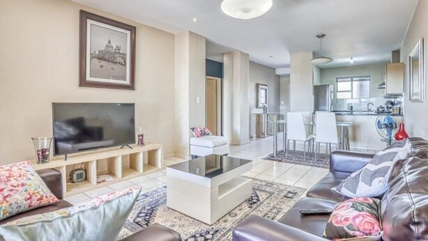 A modern living room with a flat-screen TV, a sofa, a coffee table, and a dining area with chairs and a table. - Hideout Hotel Apartment (Sandton)