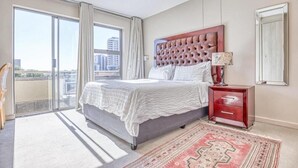 A bedroom with a large bed, a nightstand, a mirror, and a view of the city through a sliding glass door. - Hideout Hotel Apartment (Sandton)