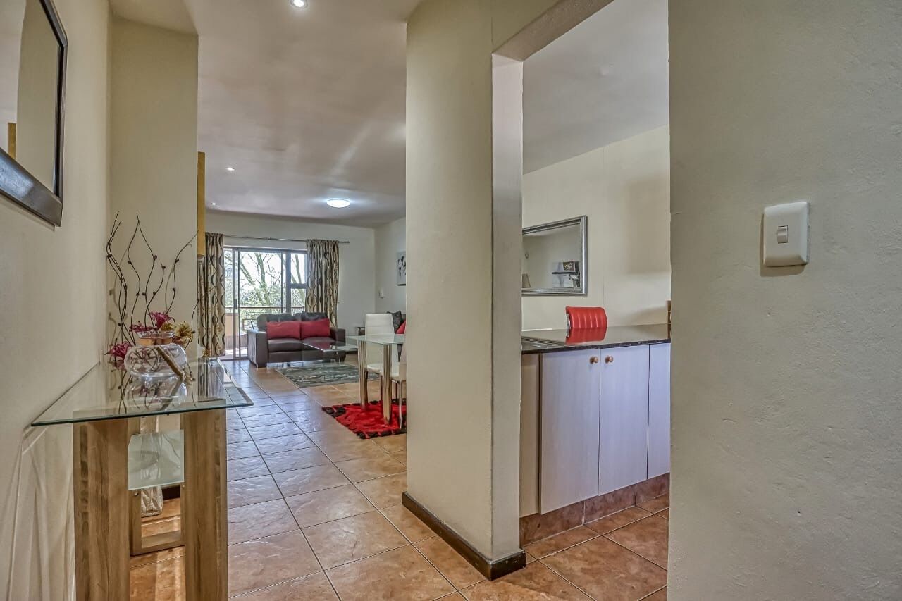 A modern interior with a glass-top dining table, a kitchen area with white cabinets, and a living space with a sofa and a red rug.