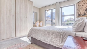 A bedroom with a bed, a nightstand, a wardrobe, and a view of buildings through a large window. - Hideout Hotel Apartment (Sandton)