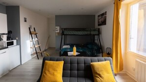 Ein kleines Zimmer mit einem Etagenbett, einer Küchenzeile, einem Sofa mit gelben Kissen und einem Fenster mit gelben Vorhängen.