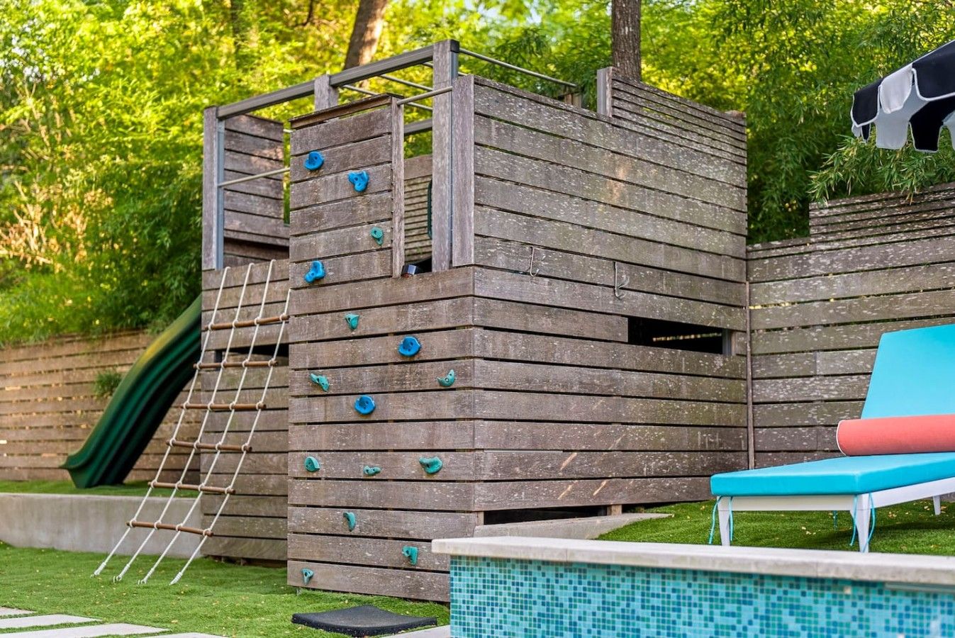 A wooden climbing structure with a ladder and climbing holds, adjacent to a pool with a blue edge and a red lounge chair.