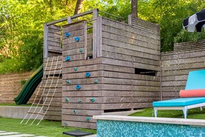 A wooden climbing structure with a ladder and climbing holds, adjacent to a pool with a blue edge and a red lounge chair. - MidMod Bungalow 5BR Heated Pool (Austin)