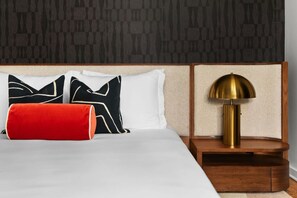 Ein Bett mit weißer Bettwäsche, einem roten Kopfkissen und zwei schwarzen Kissen mit weißen Mustern. Neben dem Bett steht ein Nachttisch aus Holz mit einer Messinglampe.