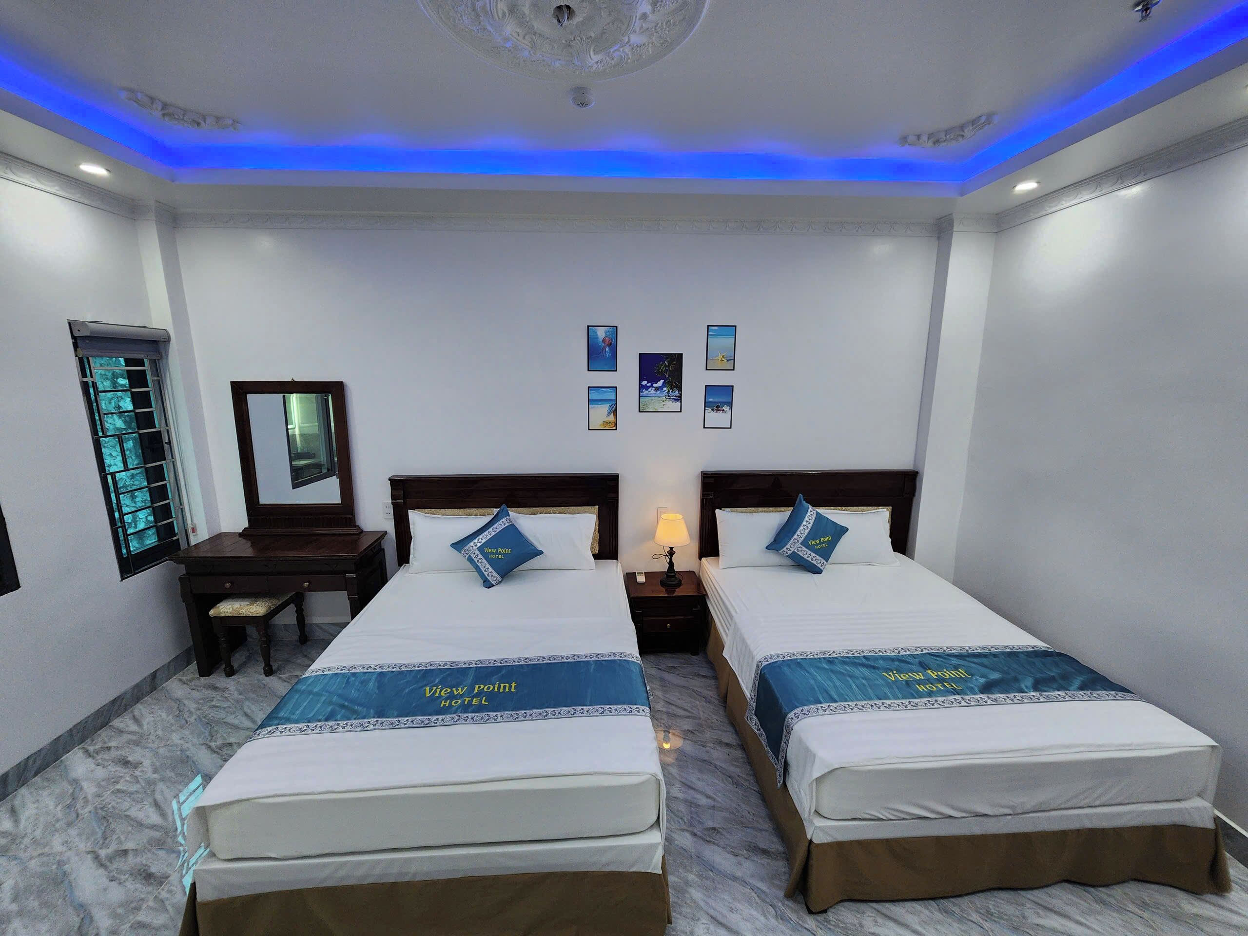 Habitación de hotel con dos camas, una mesita de noche con lámpara, cómoda y ventana con barrotes, además de cuadros en la pared.