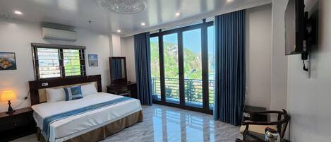 Habitación de hotel con una cama grande, un escritorio, una silla, un televisor y un balcón con vistas a zonas verdes.