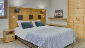 A bedroom with a wooden headboard, a bed with white bedding, a nightstand, a shelf with artwork, and a view of the outside. - Apartment with wellness area in Les Saisies (Les Saisies)