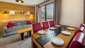 A dining area with a wooden table set for four, red chairs, and a sofa with a mirror and wall-mounted lights. - Apartment with wellness area in Les Saisies (Les Saisies)