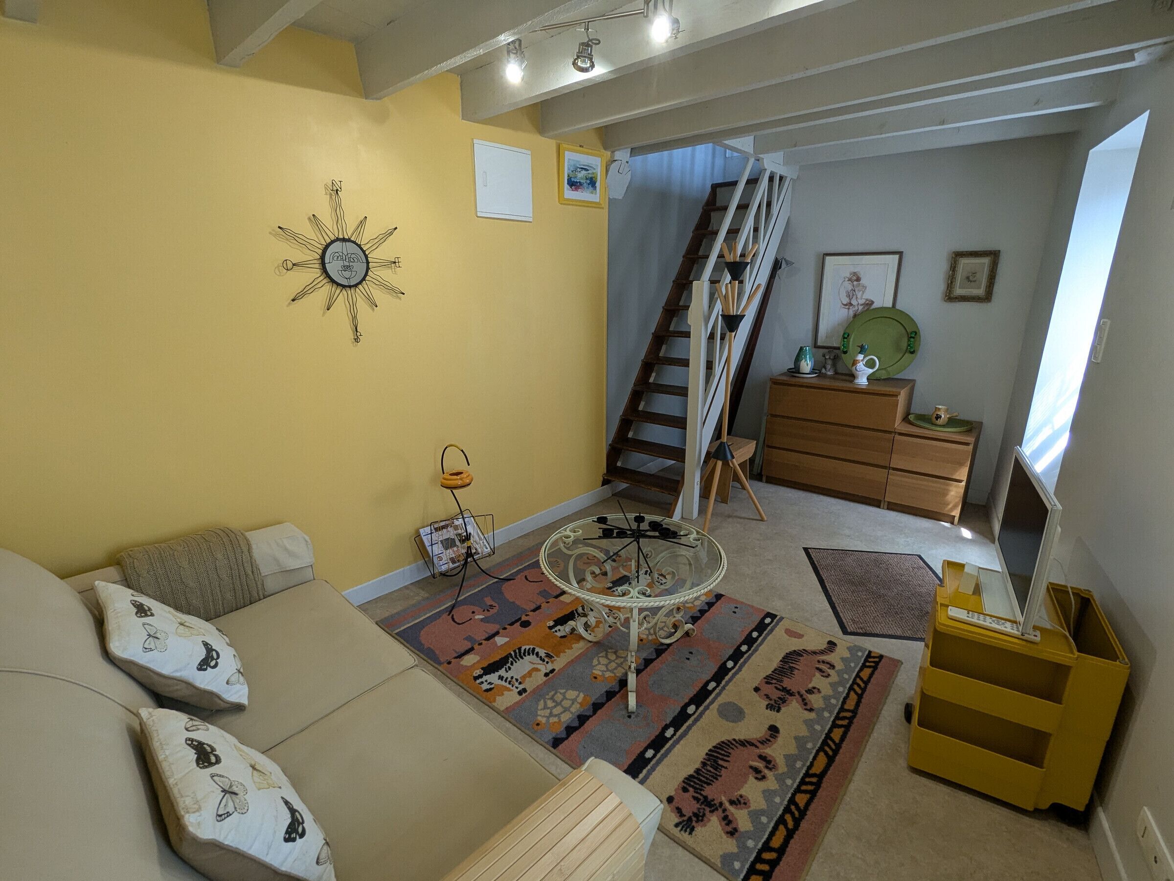 Una sala de estar con un sofá, una mesa de centro, un tapete estampado, una escalera, un aparador y una mesita con una lámpara.