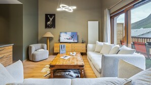 Ein modernes Wohnzimmer mit Holzboden, einem großen Fenster mit Blick auf die Berge, einem Fernseher und einem Kamin.