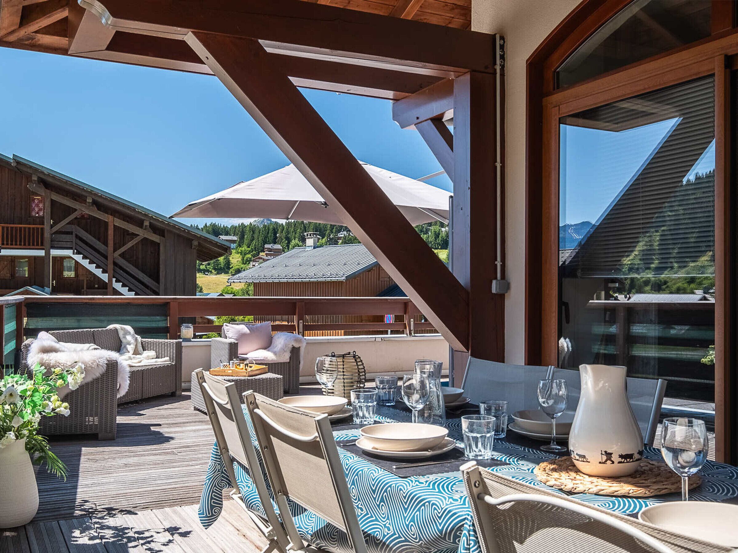 An outdoor dining area with wicker furniture, a table set for two, and a view of the surrounding buildings and mountains.