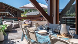 An outdoor dining area with wicker furniture, a table set for two, and a view of the surrounding buildings and mountains. - Spacious loft with spa, pool, gym, and parking (Les Saisies)