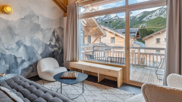 Une pièce chaleureuse avec une grande fenêtre, des poutres en bois, un jeu d’échecs et une fresque représentant une montagne.