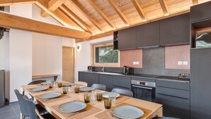Une cuisine moderne avec des poutres en bois, une grande table à manger pouvant accueillir six personnes et des meubles de rangement aux couleurs sombres.