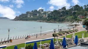 Une promenade en bord de mer offrant une vue sur l’océan, agrémentée de bancs, et une rangée de tables et de chaises en plein air, protégées par des parasols.