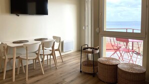 Une pièce comprenant un coin repas, un téléviseur fixé au mur et une grande fenêtre donnant sur la mer.