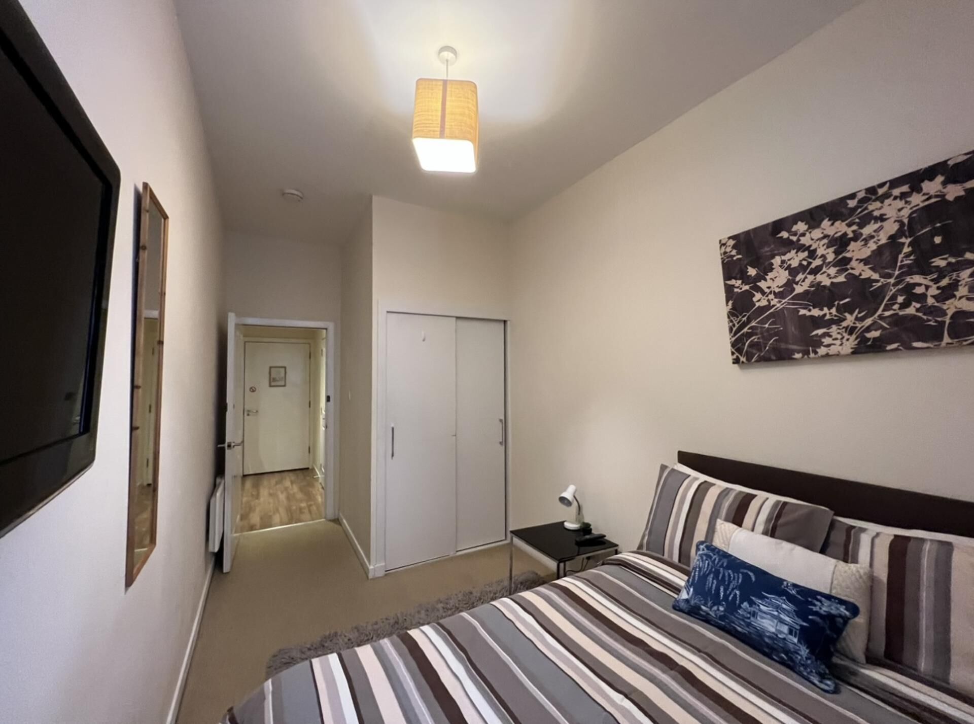 A bedroom with a bed, a nightstand, a lamp, a door, and a wall-mounted TV.
