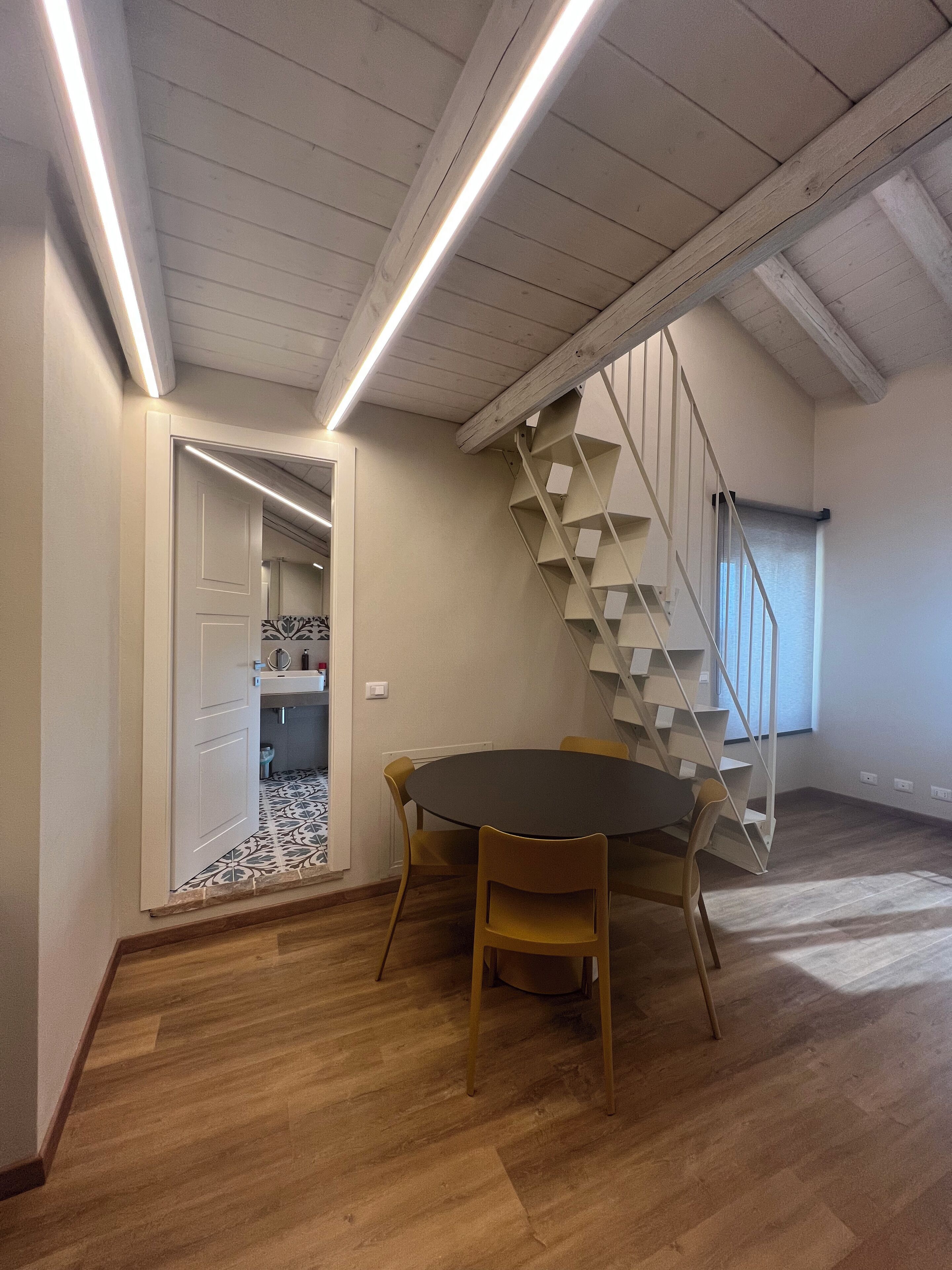 Ein Zimmer mit Holzboden, einem runden Tisch mit Stühlen, einer Treppe mit weißer Geländer und einer Tür zu einem anderen Raum.