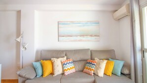 A living room with a sofa, colorful cushions, a painting on the wall, and an air conditioning unit. - Penthouse Kalymnos (Platja de L'arenal)