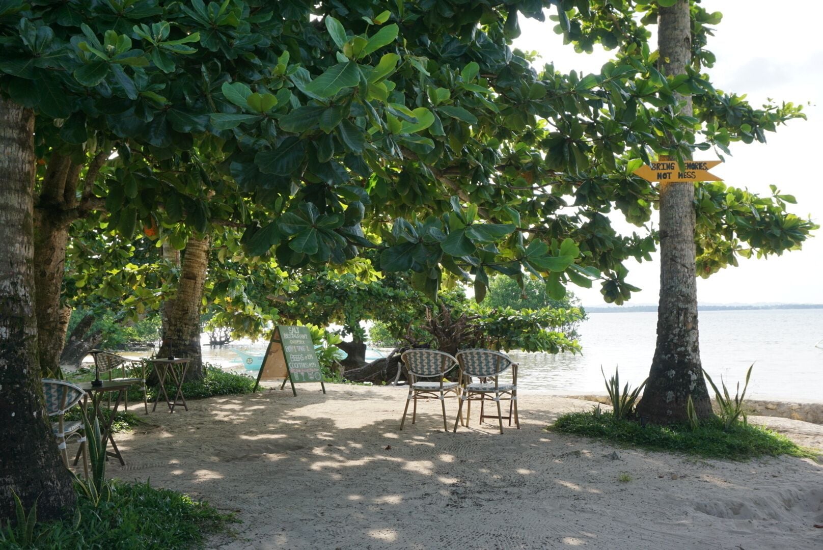 A beachside seating area with chairs and tables under trees, a sign indicating a restaurant, and a view of the sea.