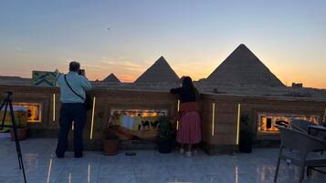 A rooftop terrace with a view of pyramids at sunset, a mural, and potted plants.