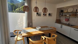A modern kitchen with a wooden table and chairs, pendant lights, and a view of a mountainous landscape through a large window. - NEXT HOUSE, FURNAS, Vila center (Furnas)