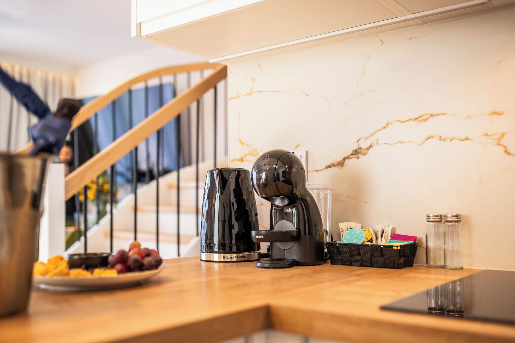 A kitchen with a coffee maker, a kettle, a plate of fruit, and a basket of keys on the counter.