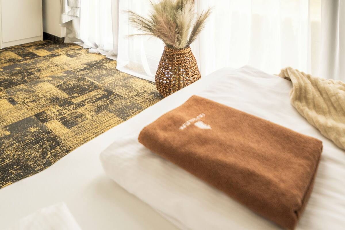 A hotel room with a bed, a window with sheer curtains, a vase with dried plants, and a patterned carpet.