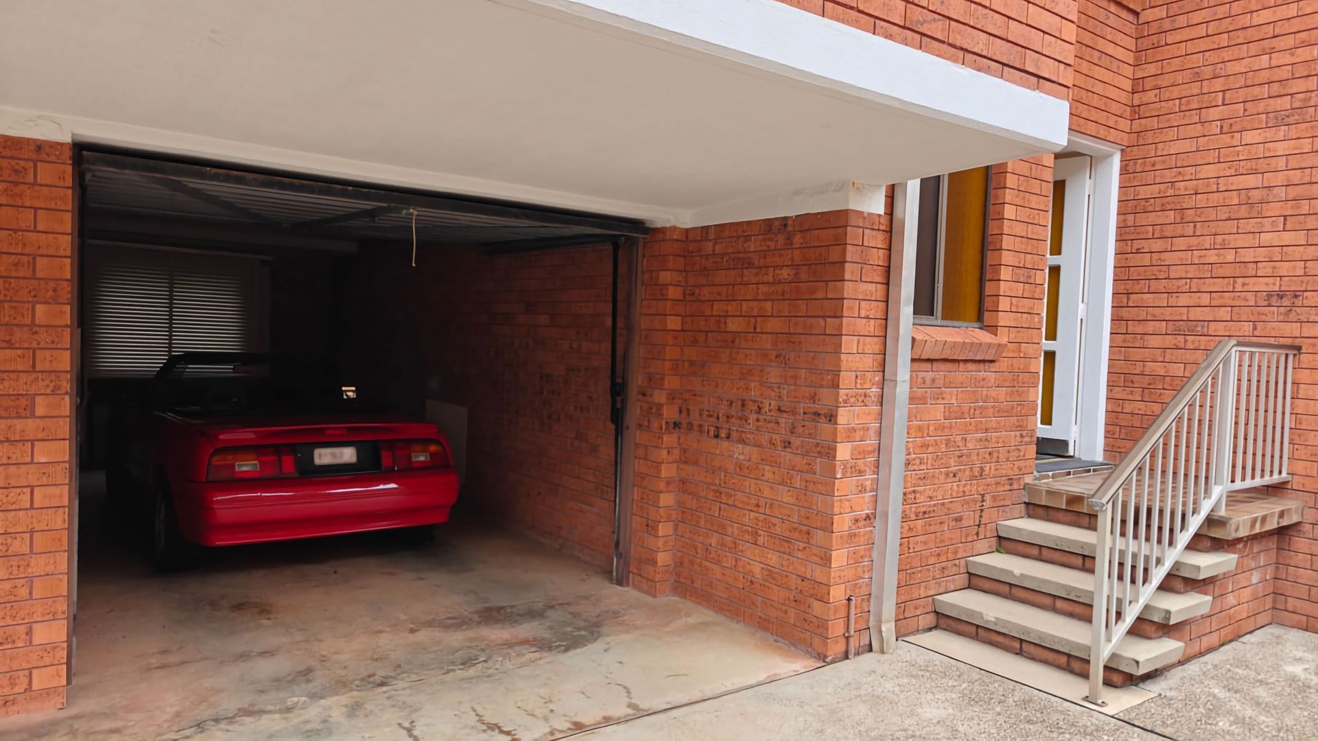 En röd bil står parkerad inuti ett garage som är anslutet till en byggnad av tegel, med ett vitt markis och en trappa av betong som leder upp till en dörr.