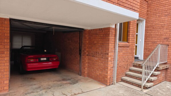 A red car parked inside a garage attached to a brick building with a white awning and a set of concrete stairs leading to a door.