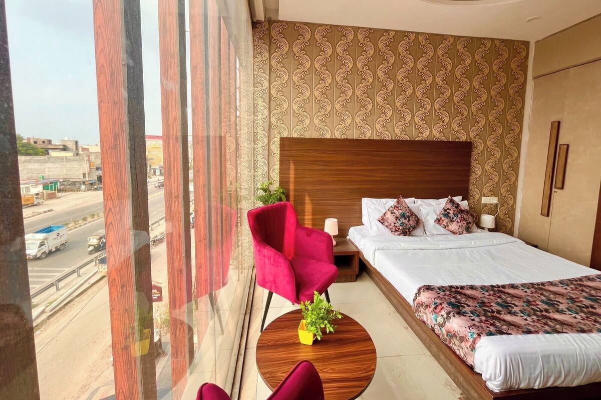 Une chambre d’hôtel avec une grande fenêtre, un lit recouvert d’une literie à motifs floraux, un fauteuil rouge et une petite table ronde sur laquelle est posée une plante.