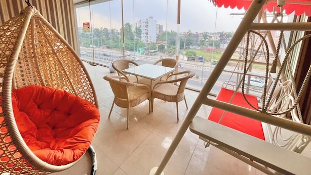A room with a hanging chair, a table, and chairs, with a view of a cityscape through large windows. - Hotel Pearl Grand by Elena Group (Dera Bassi)