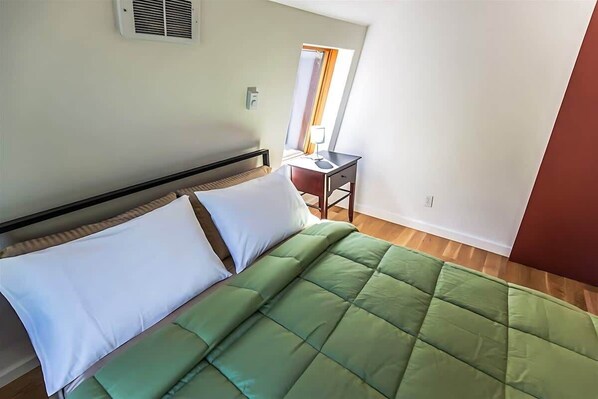 A single bed with a green quilt, white pillows, and a wooden headboard. There is a small table with a lamp and a window with curtains. - Woodland Studio Near Olympic Park, Balcony (Sequim)