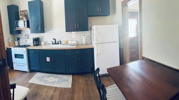 A compact kitchen with dark cabinets, a white refrigerator, a microwave, a stove, and a coffee maker. There is a wooden dining table and two chairs.