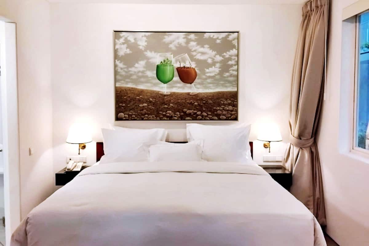 A hotel room with a large bed, two bedside tables with lamps, a ceiling fan, a window with curtains, and a wall-mounted artwork depicting plants.