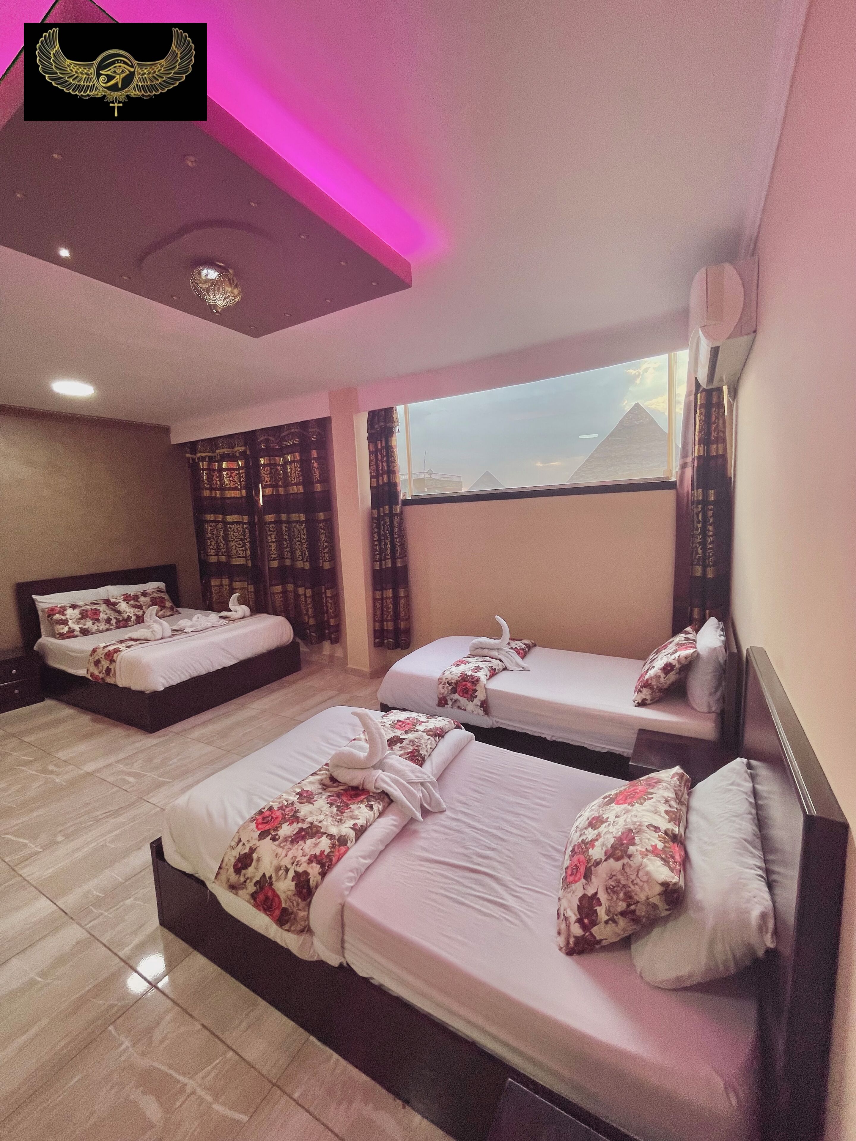 Habitación de hotel con tres camas, cada una con ropa de cama y toallas florales. La habitación tiene un ventanal con vistas a una pirámide, y el techo presenta un emblema decorativo.