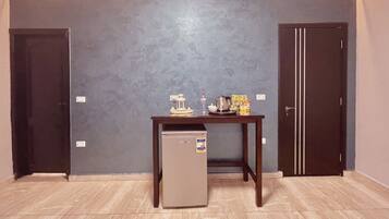 A small room with a refrigerator, a table with a kettle, a water bottle, and a box of tissues. There are two doors and a light strip along the ceiling.