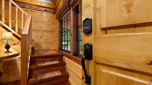 Interior de una cabaña de madera con una escalera, una puerta de madera y un ojo de cerradura, y una ventana con vistas a la vegetación.