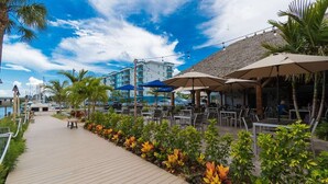 Een houten looppad met terras en parasols, omringd door tropische planten en uitzicht op een gebouw met balkons.